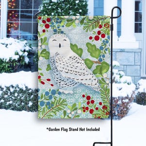 May include: A decorative garden flag featuring a snowy owl illustration. The flag has a light blue background with a pattern of green leaves, red berries, and blue berries. The owl is white with black markings.