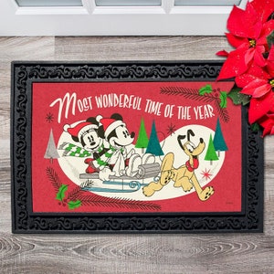 May include: A red doormat with a black border features Mickey Mouse and Minnie Mouse in winter outfits riding a sled pulled by Pluto. The text "Most Wonderful Time of the Year" is written in white on the red background.