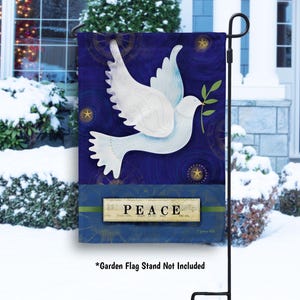 May include: A garden flag featuring a white dove carrying an olive branch against a dark blue background with gold stars. The word "PEACE" is displayed on a rectangular banner at the bottom. The flag is displayed outdoors.
