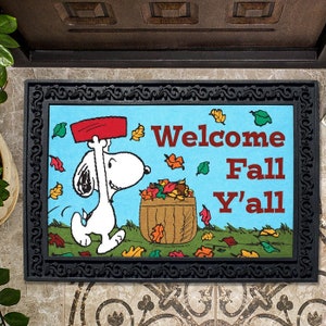 May include: A welcome mat with a blue background featuring Snoopy raking leaves. The text "Welcome Fall Y'all" is printed in red and white.