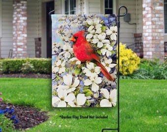 May include: A red cardinal bird perched on a branch with white dogwood blossoms. The bird is surrounded by white flowers and green leaves. The background is a blue sky. *Garden Flag Stand Not Included*