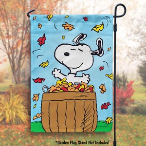May include: A garden flag featuring Snoopy, the beloved beagle from Peanuts, jumping into a pile of autumn leaves. The flag is blue with a white border and features a cartoon illustration of Snoopy with his signature smile. The flag is perfect for adding a touch of fall cheer to your outdoor space.