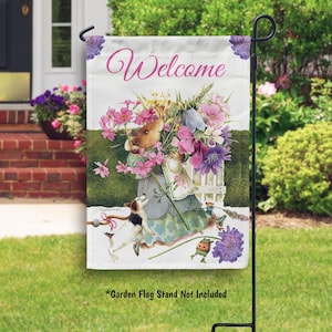 May include: A garden flag with a white background and a colorful illustration of a mouse carrying a basket of flowers. The text "Welcome" is printed in pink letters at the top of the flag. The flag is hanging from a black metal stand.