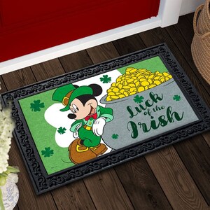 Op de afbeelding: Een groene deurmat met een zwarte rand toont Mickey Mouse gekleed in een groen kabouterkostuum dat een pot met gouden munten vasthoudt. De tekst "Luck of the Irish" staat in het groen op de deurmat.