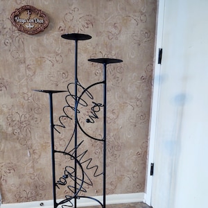 May include: Black metal candle holder with three tiers and a decorative design. The design features a circle with a wavy line and the word "Dios" in Spanish.