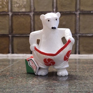 Pode incluir: Uma figura de urso polar branco segurando suas calças, que são brancas com detalhes vermelhos e desenhos florais vermelhos. Uma pequena caixa de presente verde com o texto "FOR DAD 1989" está aos pés do urso.