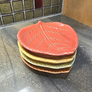 Puede incluir: Una pila de cuatro platos con forma de hoja en tonos naranja, amarillo y verde. El plato superior es de un naranja vibrante con patrones de venas detallados. Los platos están apilados sobre una encimera gris.