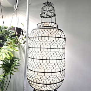 May include: A decorative black metal lantern with a geometric wire frame and a white interior. The lantern is suspended from a curved metal hook, with plants visible in the background. The lantern's design features a layered top and a circular base.