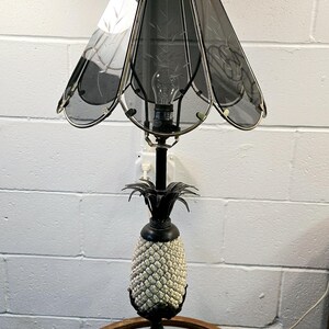 May include: A decorative table lamp with a pineapple-shaped base and a dark gray shade. The base is black and gold, with a textured, off-white pineapple design. The shade has a floral pattern and a gold trim.