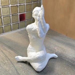 May include: A white sculpture of a person in a seated yoga pose, with hands clasped in prayer over the face. The figure is on a grey surface, with a mosaic tile wall in the background.