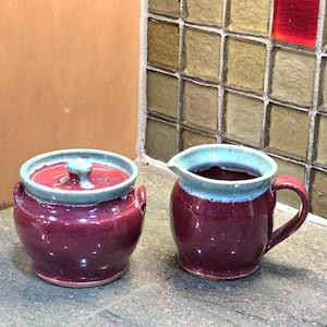 May include: A set of two ceramic sugar and creamer containers. The containers are a deep burgundy color with a light blue rim. The creamer has a handle.