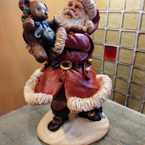 May include: A ceramic figurine of Santa Claus holding a brown teddy bear. Santa is wearing a red coat with white fur trim, a white beard, and a black belt. The figurine is standing on a white base with the words "Joy & Peace" written on it.