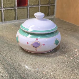 May include: A white ceramic sugar bowl with a lid. The bowl has a rounded shape with a green band around the middle and a small knob on top. It is decorated with purple and green diamond shapes and pink dots.