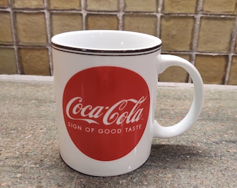 Op de afbeelding: Een witte keramische mok met een rood Coca-Cola-logo en de tekst "Sign of Good Taste".