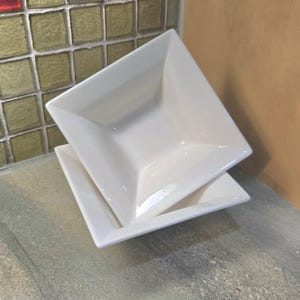 May include: Two white square ceramic bowls stacked. The bowls have a simple, modern design with slightly raised edges. The background features a mosaic tile wall and a neutral-coloured wall.