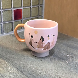May include: A pink ceramic mug with a gold handle and a gray base. The mug features a design of people holding hands and a yellow sun. The mug is sitting on a gray countertop.
