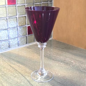 Könnte beinhalten: Ein Weinglas mit einer tiefvioletten Schale und einem klaren Stiel. Das Glas hat eine klassische Form mit einer breiten Oberseite, die sich zu einer schmalen Basis verjüngt. Das Glas steht auf einer hellen Oberfläche.