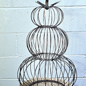 May include: A decorative, three-tiered pumpkin made of dark brown metal wire. The pumpkin has a stem and is sitting on a woven, circular base. The pumpkin is a fall-themed decoration.