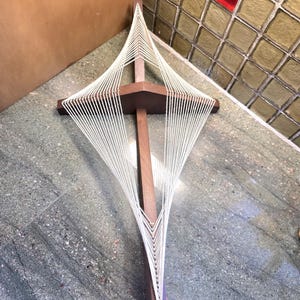 May include: A dark brown wooden cross with a geometric design made of white string. The cross has a vertical post and a horizontal beam. The string is strung in a diamond pattern. The cross is displayed on a gray and green tiled surface. The cross is a decorative item.