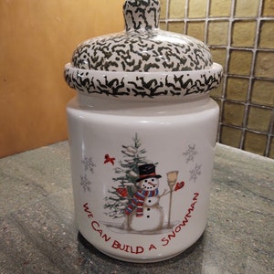Pode incluir: Jarro de biscoitos de cerâmica branca com uma tampa salpicada de verde e preto. O jarro apresenta um boneco de neve com um cachecol e um chapéu vermelhos, segurando uma vassoura. O texto "We can build a snowman" está escrito em torno do boneco de neve.