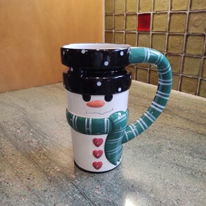 Könnte beinhalten: Eine weiße Keramik-Schneemann-Tasse mit einem schwarzen Hut, einem grünen Schal und roten Herzen. Die Tasse hat einen Henkel in Form eines grün-weiß gestreiften Schals.
