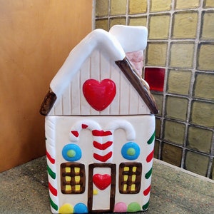 Könnte beinhalten: Ein Keramik-Lebkuchenhaus-Keksdose mit weißem Dach und roten Herzakzenten. Das Haus hat Zuckerstangendetails, bunte Punkte und einen Schornstein. Die Fenster sind braun mit gelben Innenräumen, und die Tür hat ein rotes Herz.