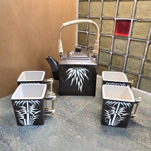 Puede incluir: Un juego de té de cerámica cuadrada que incluye una tetera marrón oscuro con asa envuelta en bambú y tres tazas cuadradas a juego. Cada pieza está decorada con un motivo de hojas de bambú pintado en blanco. El interior es blanco roto.