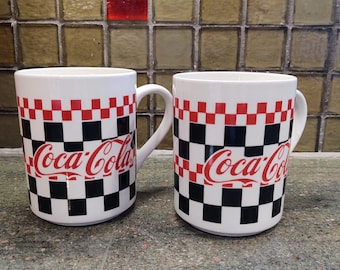 Op de afbeelding: Twee witte keramische Coca-Cola mokken. Elke mok heeft een rood, zwart en wit geblokt patroon rond de bovenkant en het Coca-Cola logo in rood. De mokken hebben een gebogen handvat.