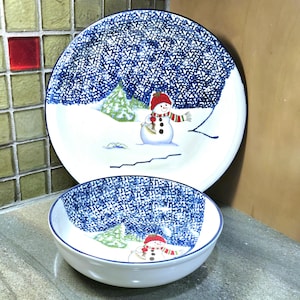 May include: A white ceramic plate and bowl set with a winter scene. The plate and bowl feature a blue speckled border and a snowman wearing a red hat and scarf. The background depicts a snowy landscape with green trees.