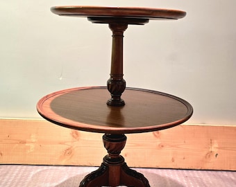 May include: A two-tiered wooden table with a round top and three legs. The table is made of dark wood and has a carved design on the legs.