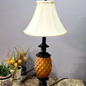 Puede incluir: Una lámpara de mesa con una base con forma de piña y una pantalla blanca. La base es de metal marrón y tiene un acabado texturizado. La pantalla es de tela y tiene forma de campana.