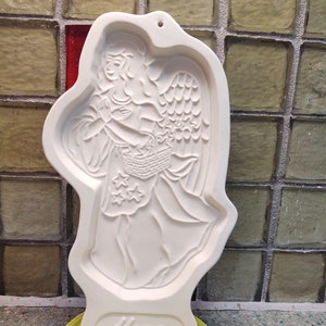 Puede incluir: Cortador de galletas de cerámica blanca con forma de ángel sosteniendo una cesta. El cortador tiene un diseño en relieve y la palabra "Hope" está impresa en la parte inferior.