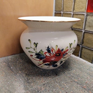 May include: White ceramic vase with a gold rim and a floral design featuring a red robin, holly, and berries.