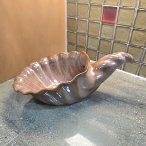 Pode incluir: Uma tigela de cerâmica em forma de concha em tons de castanho e bege. A tigela tem uma borda ondulada e texturizada e uma alça curva. O acabamento brilhante reflete a luz, destacando os detalhes do design. Esta peça decorativa é adequada para servir ou exibir.