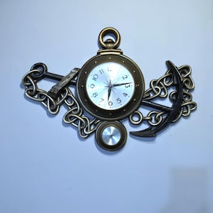 May include: A decorative wall clock with a nautical theme. The clock is designed to resemble a pocket watch, surrounded by an anchor, chain, and a small circular element. The clock face has a white background with black numerals and hands.