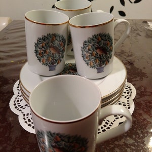 May include: Four white ceramic mugs with gold trim and a pear tree design. The mugs are arranged on a white saucer with a lace doily underneath.