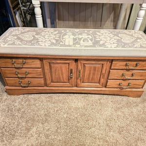May include: A wooden storage chest with a padded top featuring a floral design. The chest has four drawers and two doors with brass handles.