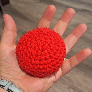 May include: A bright red, crocheted sphere, approximately 3 inches in diameter, is held in an open hand. The handmade item features a tight stitch pattern and a rounded shape. The background is a blurred wooden surface.