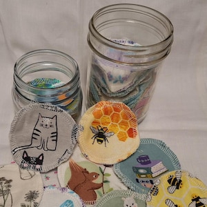 May include: Reusable cotton rounds in a glass jar with various designs including a cat, a bee, a squirrel, a butterfly, a llama, and a book. The rounds are white with colorful designs.