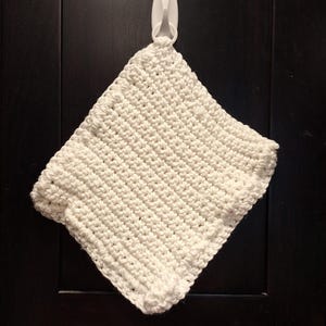 May include: A square, off-white, crocheted washcloth hanging from a white plastic hook. The washcloth has a textured surface and a thick, decorative border. The background is a dark, solid color.
