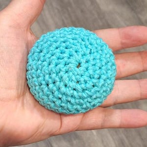 May include: A round, turquoise crocheted item. The handmade object is a solid, circular shape with a textured surface created by the visible stitches. The item is held in a hand, with the background out of focus.