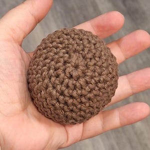 May include: A small, round, brown crocheted item, held in a hand. The item has a textured surface created by the crochet stitches. The color is a warm, earthy brown.