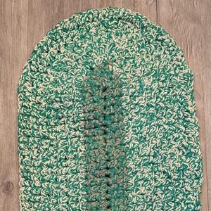 May include: Oval-shaped, hand-crocheted rug in shades of green and cream. The rug features a central stripe of a darker green color. The texture is dense and the stitches are clearly visible, suggesting a handmade item. The rug is approximately 30 inches long.