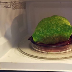 A reusable microwave popcorn bag in green paisley fabric sitting on a microwavable plate inside a microwave after the popcorn kernels inside have been popped, giving it a puffed up and full appearance, an example of result of this sewing tutorial.