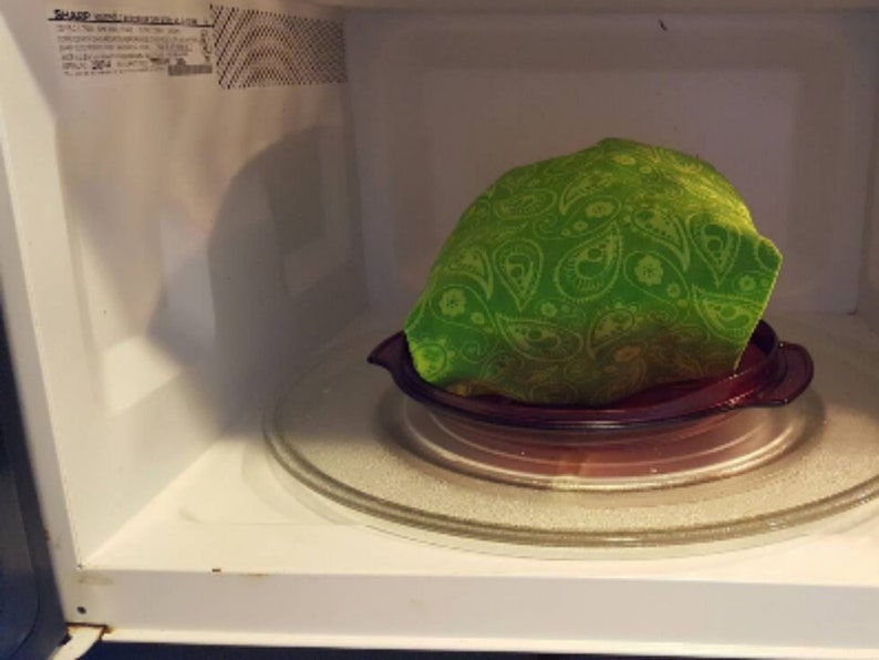 A reusable microwave popcorn bag in green paisley fabric sitting on a microwavable plate inside a microwave after the popcorn kernels inside have been popped, giving it a puffed up and full appearance, an example of result of this sewing tutorial.