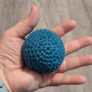 May include: A teal blue, crocheted ball. The ball is round and has a textured surface. The ball is held in an open hand. The background is a light brown color.