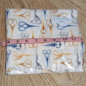 May include: A package of 24 quilt squares with a repeating pattern of scissors in blue, gold, and gray on a cream background. The squares are approximately 11 inches wide, measured with a pink tape measure.