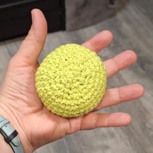May include: A small, round, crocheted ball in a bright, spring green color. The ball is held in an open hand, showcasing the texture of the yarn and the handmade quality. The item appears to be a decorative or functional craft item.