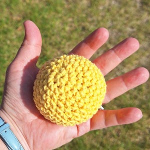 May include: A bright yellow, crocheted ball. The ball is round and has a textured surface. The image is taken outdoors with a blurred green background.
