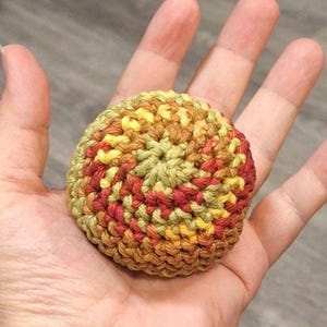 May include: A small, round, crocheted sphere featuring a variegated yarn pattern in shades of olive green, golden yellow, and deep red. The item is constructed with a tight, circular stitch pattern that radiates from the center outward.